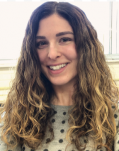Leah is smiling at the camera. She has wavy light brown hair and is wearing a gray and black polka-dot shirt. The background behind her is a white wall and light window.