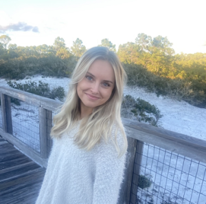 Olivia is smiling on a wooden bridge. She has long blonde hair and is wearing a white fuzzy sweater. In the background is white sand and green shrubs and trees.